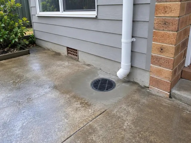 Round outdoor drain grate in a concrete driveway beside a house wall in Kingsgrove